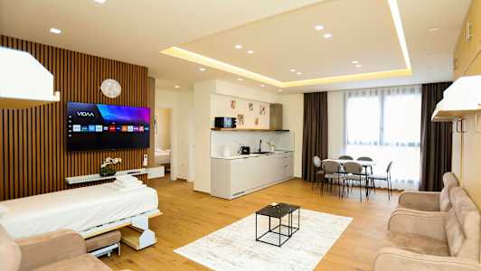 Ett modernt hotellrum med öppen planlösning inkluderande en bäddsoffa, TV, köksområde, matbord och stora fönster med gardiner. (AI-genererad text)