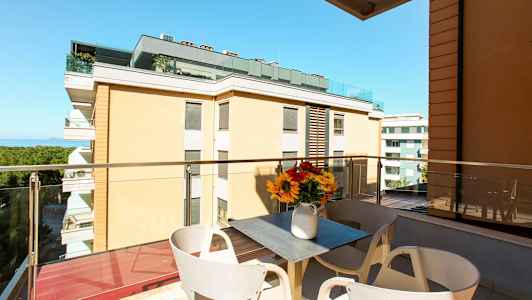 En balkong med utsikt över byggnader och havet. På balkongen finns ett bord med en vas med blommor. (AI-genererad text)