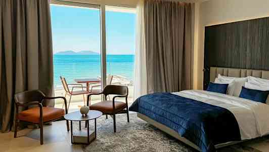 Ett hotellrum med havsutsikt, innehållande en dubbelsäng, två stolar, ett litet bord och en glasdörr till en terrass. (AI-genererad text)