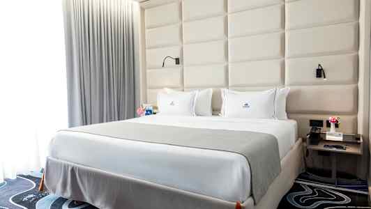 Ett sovrum med en stor säng, vita lakan och ett grått överkast. Bakom sängen finns en stoppad huvudgavel, och vid sidorna står nattduksbord med lampor. (AI-genererad text)