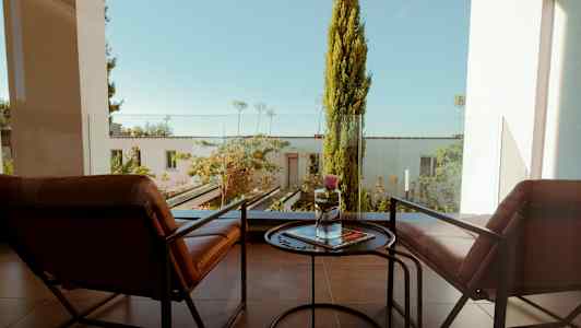 Två stolar och ett litet bord på en balkong med utsikt över en grönskande trädgård. (AI-genererad text)