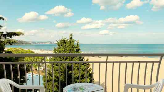 Utsikt från en balkong mot en strand och havet, med stolar och ett bord i förgrunden. (AI-genererad text)