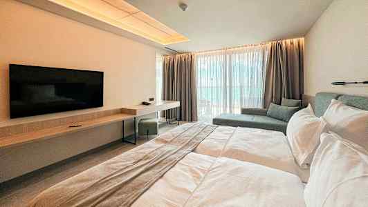 Ett modernt hotellrum med en dubbelsäng, en väggmonterad TV, skrivbord och sittområde vid stora fönster. (AI-genererad text)