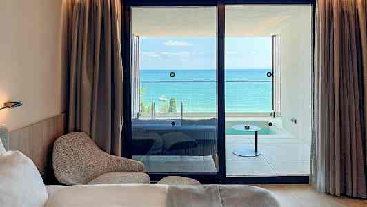 Ett hotellrum med en säng och fåtölj, samt utsikt över en balkong med havet i bakgrunden. (AI-genererad text)