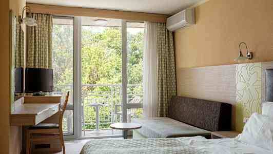 Ett hotellrum med säng, skrivbord, stol och soffa. Fönster och balkong med utsikt mot grönska. (AI-genererad text)