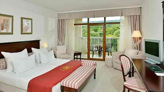 Hotellrum med säng, sittgrupp och balkong med utsikt över grönska. (AI-genererad text)