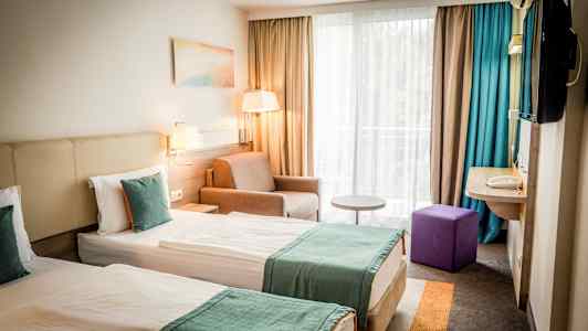 Två enkelsängar med turkosa överkast i ett hotellrum med fönster och sittgrupp. (AI-genererad text)