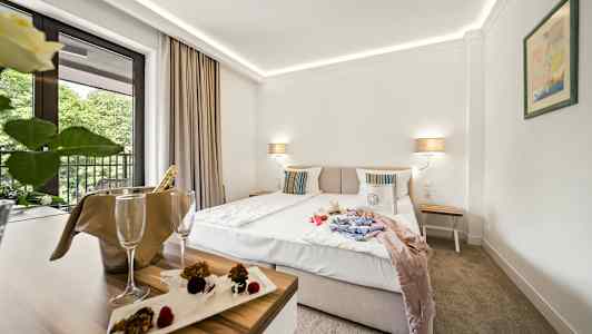 Hotellrum med en dubbelsäng, nattduksbord och balkong med utsikt mot träd. Ett bord med champagnhängare och dessert. (AI-genererad text)