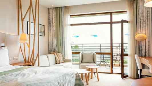 Ett hotellrum med säng, sittgrupp, bord och öppen balkongdörr. (AI-genererad text)