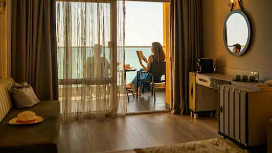 Hotellrum med en balkong där någon sitter och läser med havsutsikt. (AI-genererad text)