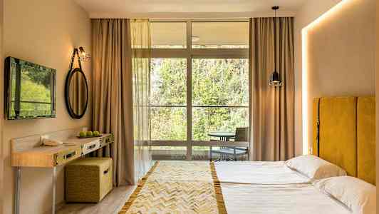 Ett hotellrum med en dubbel bädd, gul sänggavel, litet skrivbord, och en stor fönsterfront mot naturen. (AI-genererad text)