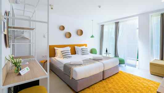 Ett ljust och modernt hotellrum med två enkelsängar, gula detaljer och stora fönster med gardiner. (AI-genererad text)