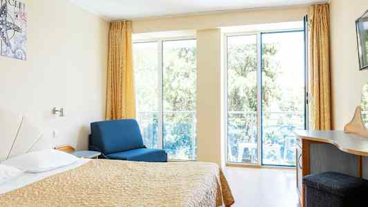 Rum med säng, blå fåtölj och skrivbord, med stora fönster mot balkong. (AI-genererad text)