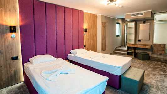 Två enkelsängar med vita lakan i ett hotellrum med lila sänggavel, träpaneler och en arbetsyta med spegel. (AI-genererad text)