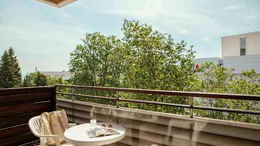 En balkong med ett bord, två plaststolar, en väska, solglasögon och en tidning, samt utsikt över träd och byggnader. (AI-genererad text)
