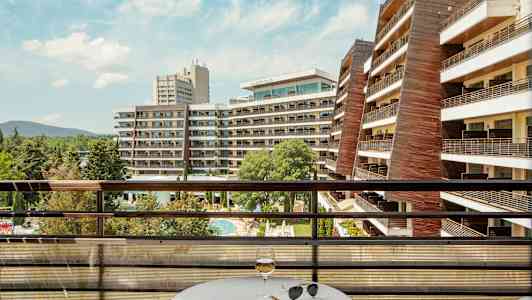 Utsikt från en balkong med bord och glas över hotellbyggnader och poolområde. (AI-genererad text)