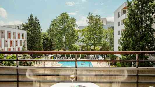 Vy från en balkong med utsikt över en pool och grönska, inkluderar ett bord med ett glas vin och en bok. (AI-genererad text)