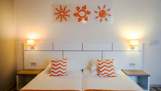 Ett hotellrum med två enkelsängar, orange kuddar och väggdekorationer i form av solar. (AI-genererad text)