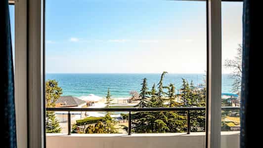 Utsikten genom ett fönster visar havet, tallar, hus och en balkong. (AI-genererad text)