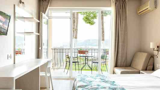 Ett hotellrum med en säng, ett bord och en stol, samt en balkong med utemöbler och utsikt mot naturen. (AI-genererad text)