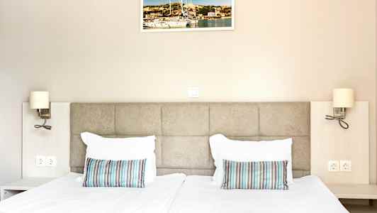 Två sängar med vit sängkläder och randiga kuddar, väggen bakom har en tavla och sänglampor. (AI-genererad text)