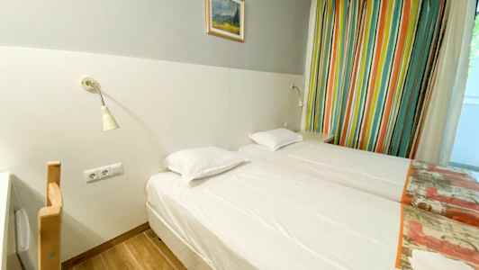 Ett hotellrum med två enkelsängar, ett ljusgrått väggfält med en målning ovanför, och färgglada gardiner vid sidan. (AI-genererad text)