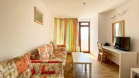 Bild av ett vardagsrum med soffa, soffbord, en TV och ett skrivbord. En dörr och ett fönster med färgglada gardiner är synliga. (AI-genererad text)