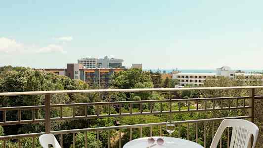 Utsikt från en balkong med bord och stol, med grönska, byggnader och hav i bakgrunden. (AI-genererad text)