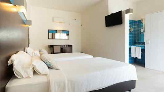 Ett hotellrum med två sängar, vita lakan och en väggmonterad TV. Spegel och skrivbord finns mot ena väggen, samt ett öppet badrum med blå kakel. (AI-genererad text)