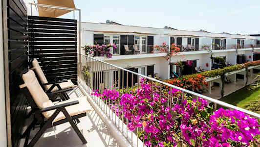 Balkong med utomhusstolar och blommande växter framför en byggnad med flera lägenheter. (AI-genererad text)