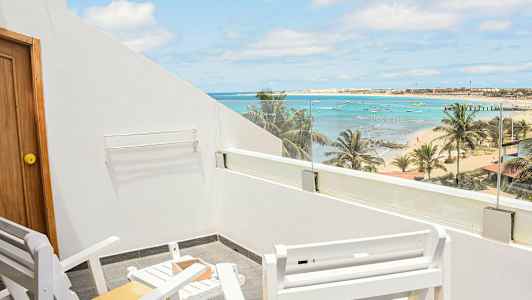 Bilden visar en uteplats med vita stolar och bord samt utsikt över en strand med palmer, hav och små båtar. (AI-genererad text)