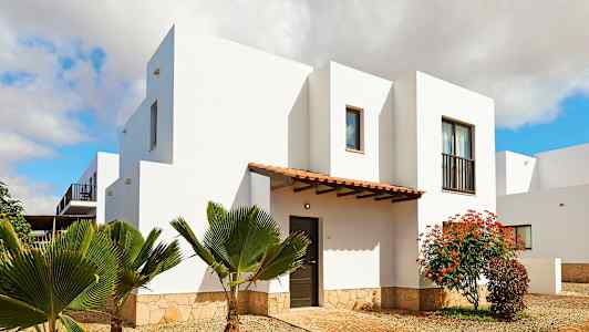 En vit byggnad med en stenbas, omgiven av växter och en träingång. (AI-genererad text)