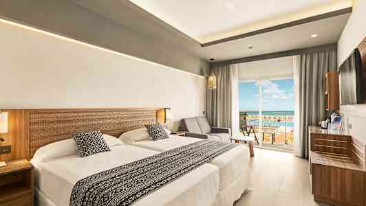 Ett hotellrum med två sängar och en sittgrupp bredvid en glasdörr som leder till en balkong med utsikt. (AI-genererad text)