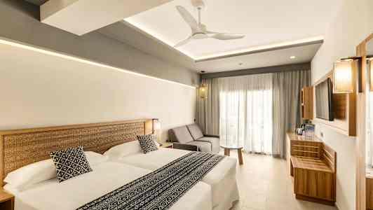 Ett modernt hotellrum med två sängar, en soffa och ett bord. Stilen är ljus och minimalistisk. (AI-genererad text)