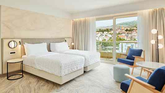 Hotellrum med två enkelsängar, sittgrupp och balkong med utsikt över en stad. (AI-genererad text)