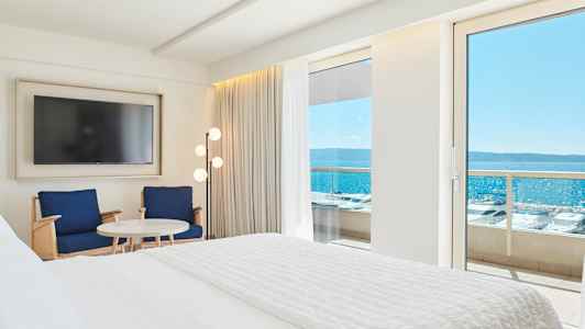 Interiör av ett hotellrum med en stor säng, två stolar, ett bord och en tv. Utsikt över havet genom en balkongdörr. (AI-genererad text)