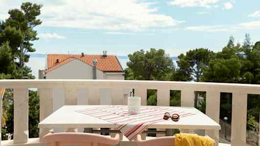 En balkong med bord täckt av randig duk, solglasögon och gul sjal, med utsikt mot havet och hus. (AI-genererad text)