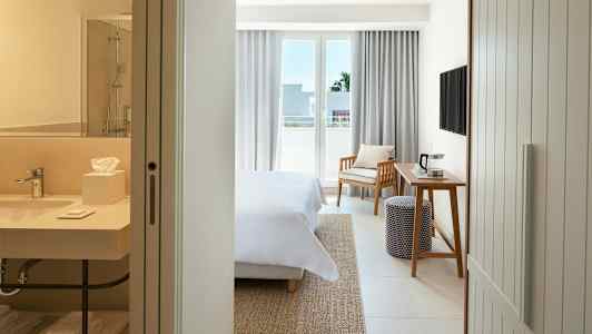 Ett hotellrum med en säng, ett skrivbord, en stol och en balkong. I förgrunden syns ett badrum. (AI-genererad text)