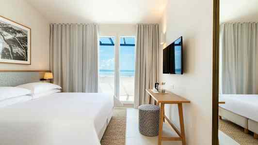 Ett hotellrum med en stor säng, ett skrivbord, och en TV på väggen. Stora fönster med utsikt mot havet. (AI-genererad text)