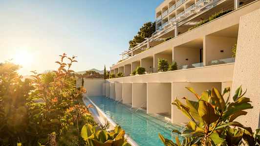 En hotellbyggnad med en längsgående pool framför, omgiven av växter och soluppgång. (AI-genererad text)