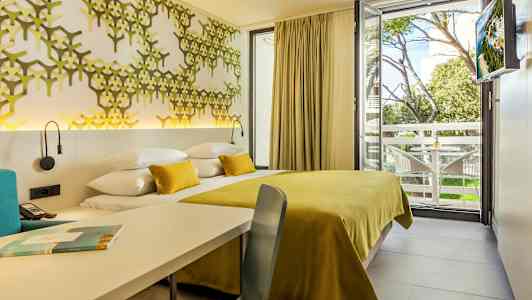 Fotografi av ett hotellrum med en gul sängöverkast och dekorativ väggpanel samt utsikt mot en trädkant och balkong. (AI-genererad text)