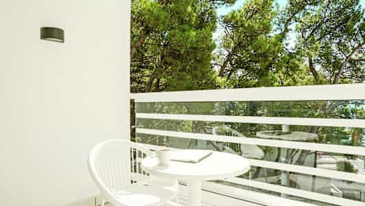 En balkong med ett vitt runt bord och en vit stol. Utsikt över gröna träd. (AI-genererad text)