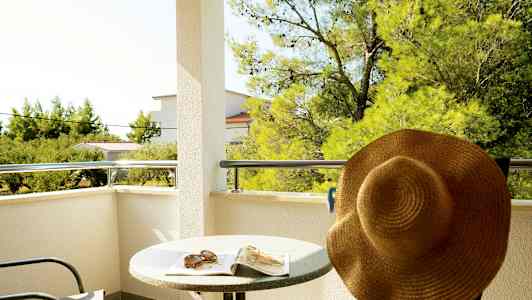 Balkong med bord, stol och solhatt, med utsikt över träd och byggnader. (AI-genererad text)