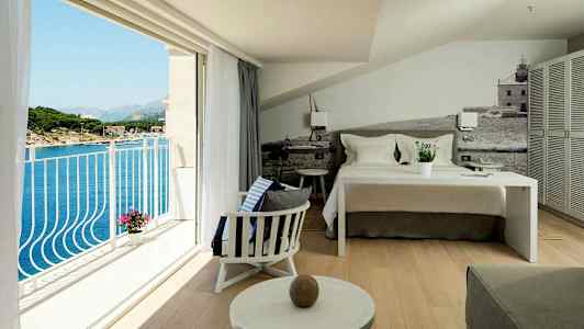 En hotellrum med en bäddad säng, sittområde och en balkong med utsikt över havet. (AI-genererad text)