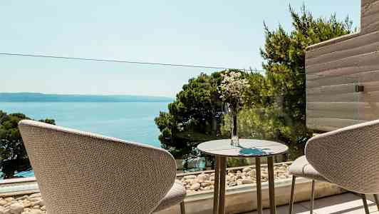 Två stolar står vid ett bord med en vas med blommor, med utsikt över havet och grönskande träd. (AI-genererad text)