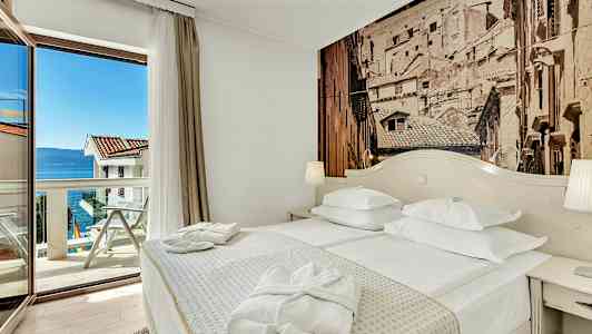 Ett hotellrum med en dubbelsäng, ett bord, och en glasdörr som leder till en balkong med utsikt över havet. (AI-genererad text)