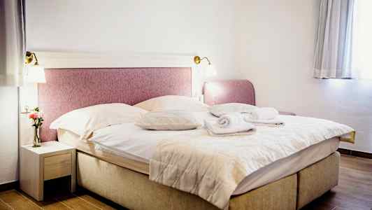 Ett rum med en dubbelsäng bäddad med filtar och kuddar, två uppvikta handdukar ovanpå. Ett nattduksbord med en vas och en blomma till vänster och fönster med gardiner till höger. (AI-genererad text)