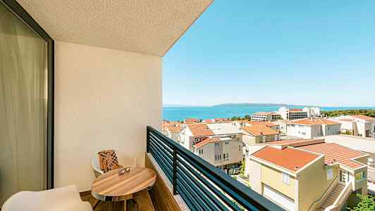 Utsikt från en balkong med två stolar, ett bord och utsikt över en stadsbebyggelse och havet. (AI-genererad text)