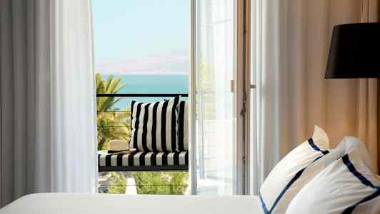 Säng med vita och blå lakan, bredvid en balkong med utsikt över havet. (AI-genererad text)