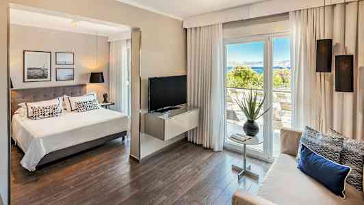 Interiör av ett hotellrum med sittgrupp, säng och utsikt mot en terrass med hav i bakgrunden. (AI-genererad text)